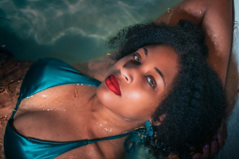 A photo from the chest up of an attractive woman with red lipstick and a bikini top laying n in water with a seductive expression.